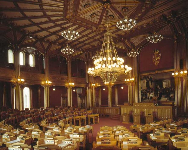 Bilde av en staselig sal med mørke trepaneler, høyt dekorert tak, flere lysekroner og rekker av pulter og stoler, som minner om et parlament eller bystyresal.