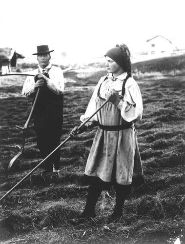 Historisk svart-hvitt-foto av en kvinne og mann i tradisjonelle arbeidsklær som arbeider med høy på et jorde ved hjelp av river og annet jordbruksutstyr.