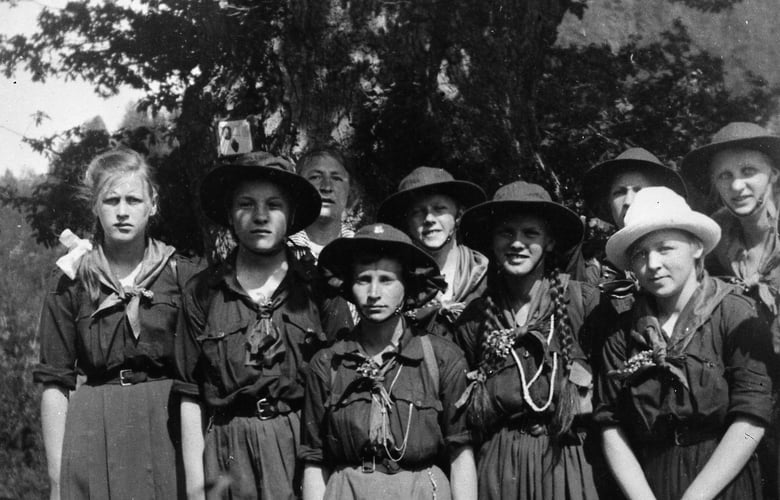 Historisk svart-hvitt gruppefoto av speiderjenter i uniform og hatter, fotografert utendørs foran et stort tre.