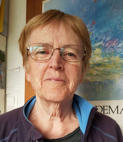 Et portrettfoto av en kvinne med briller, tatt innendørs foran en fargerik plakat eller et maleri.