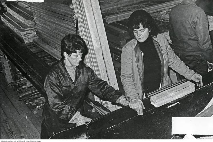 To kvinner arbeider med å skjære til plank i fabrikklokale. Mann i bakgrunnen.