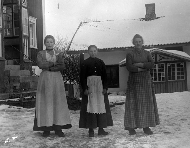 Historisk fotografi av tre kvinner i forklær som står utendørs i snødekt gårdstun.