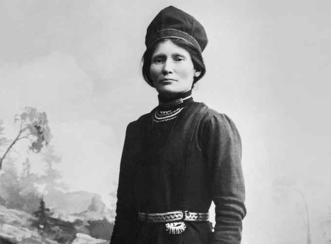 Svart-hvitt bilde av kvinne med tradisjonelle samiske klær og hodeplagg.