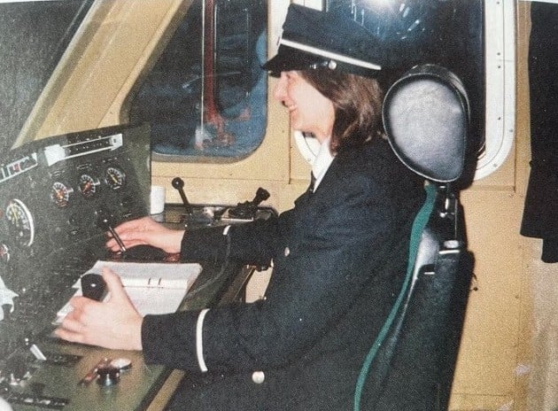 Kvinne sitter ved kontrollpanelet i førerhuset på et tog eller fartøy, iført uniform.