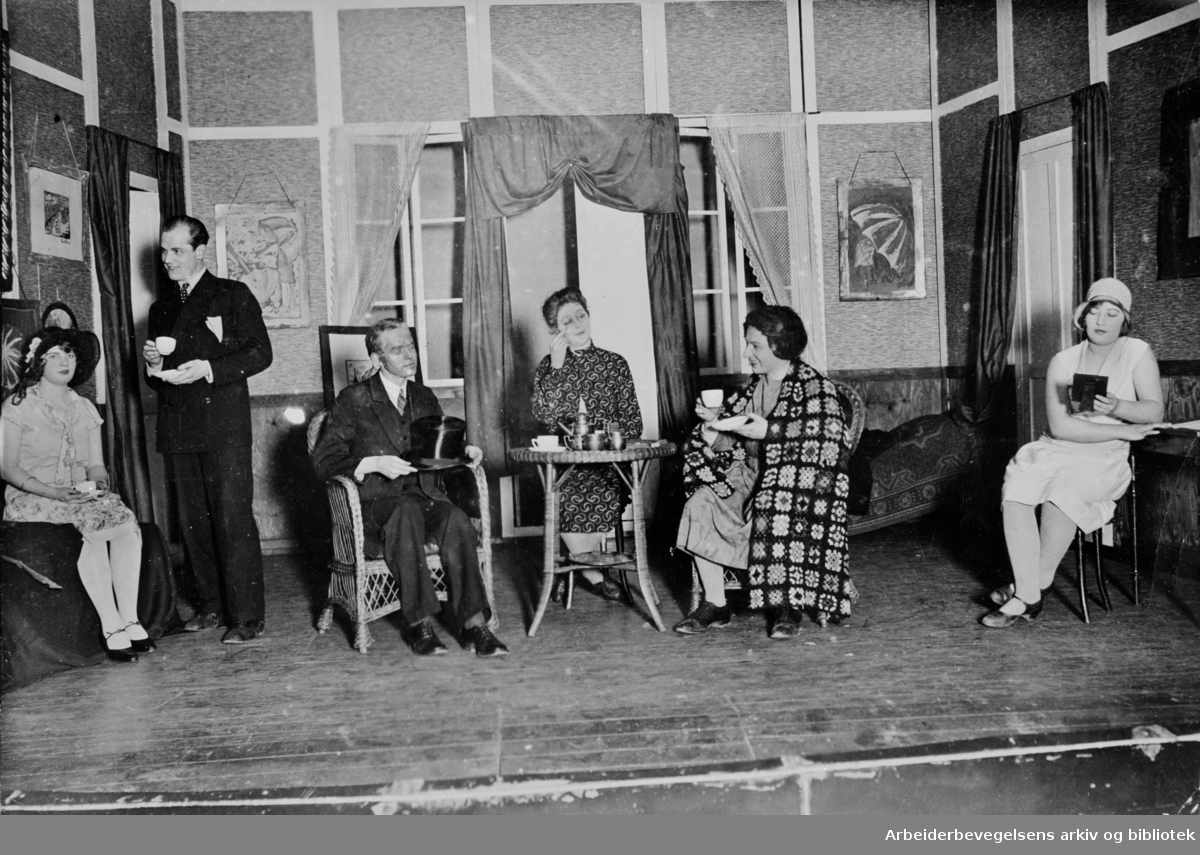 Teaterscene med 6 skuesplillere, kvinner og menn. Kulisser fra en dagligstue der det drikkes kaffe eller te.