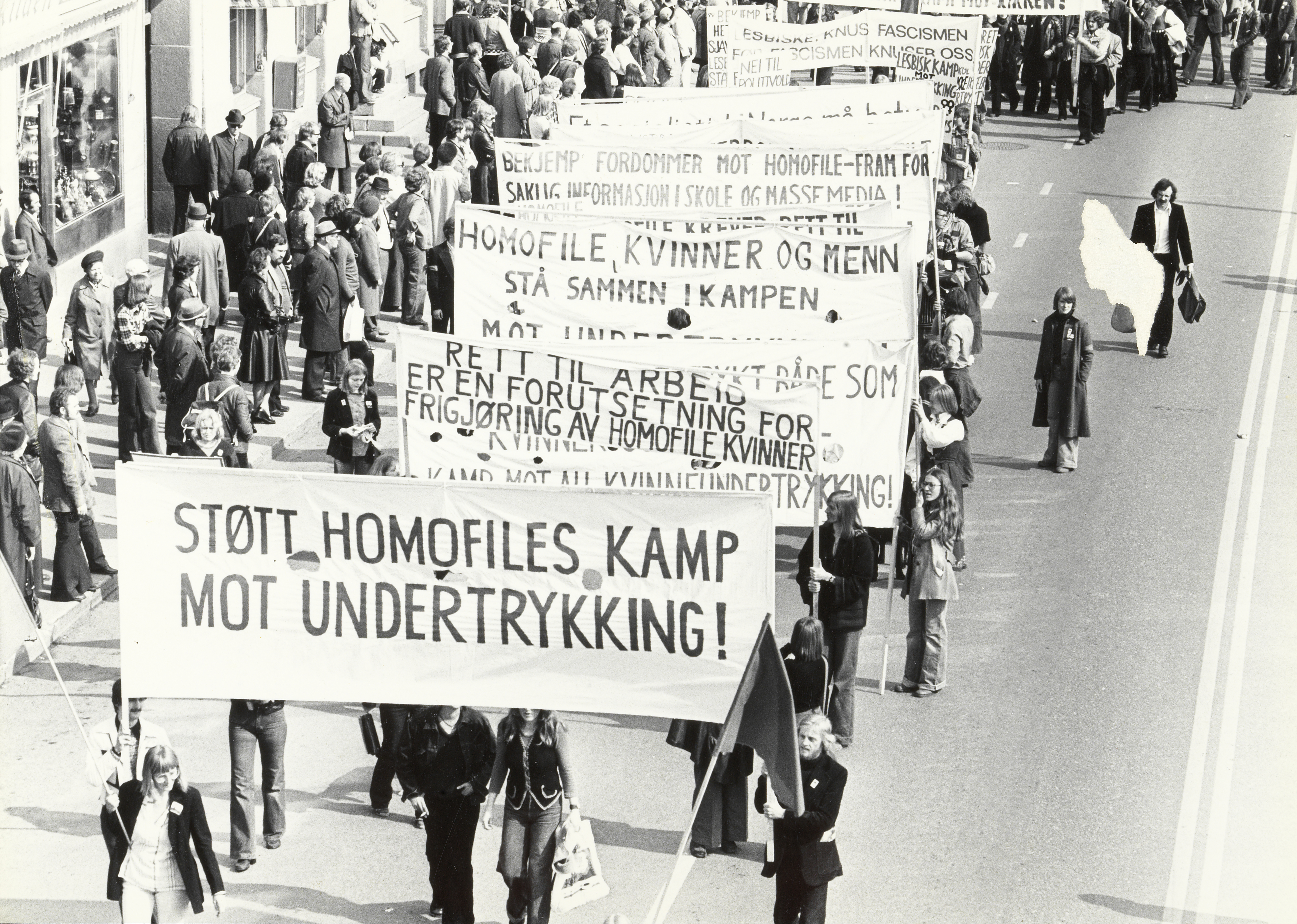 Demonstrasjon, ukjent år, mest sannsynlig 1970-årene. Paroler: Støtt homofiles kamp mot undertrykking! Rett til arbeid er en forutsetning for frigjøring av homofile kvinner. Homofile kvinner og menn - stå sammen i kampen mot undertrykking. Bekjemp fordommer mot homofile - fram for saklig informasjon i skole og massemedia! … Knus fascismen før fascismen knuser oss. Plakat med Nei til politivold og Lesbisk kamp…