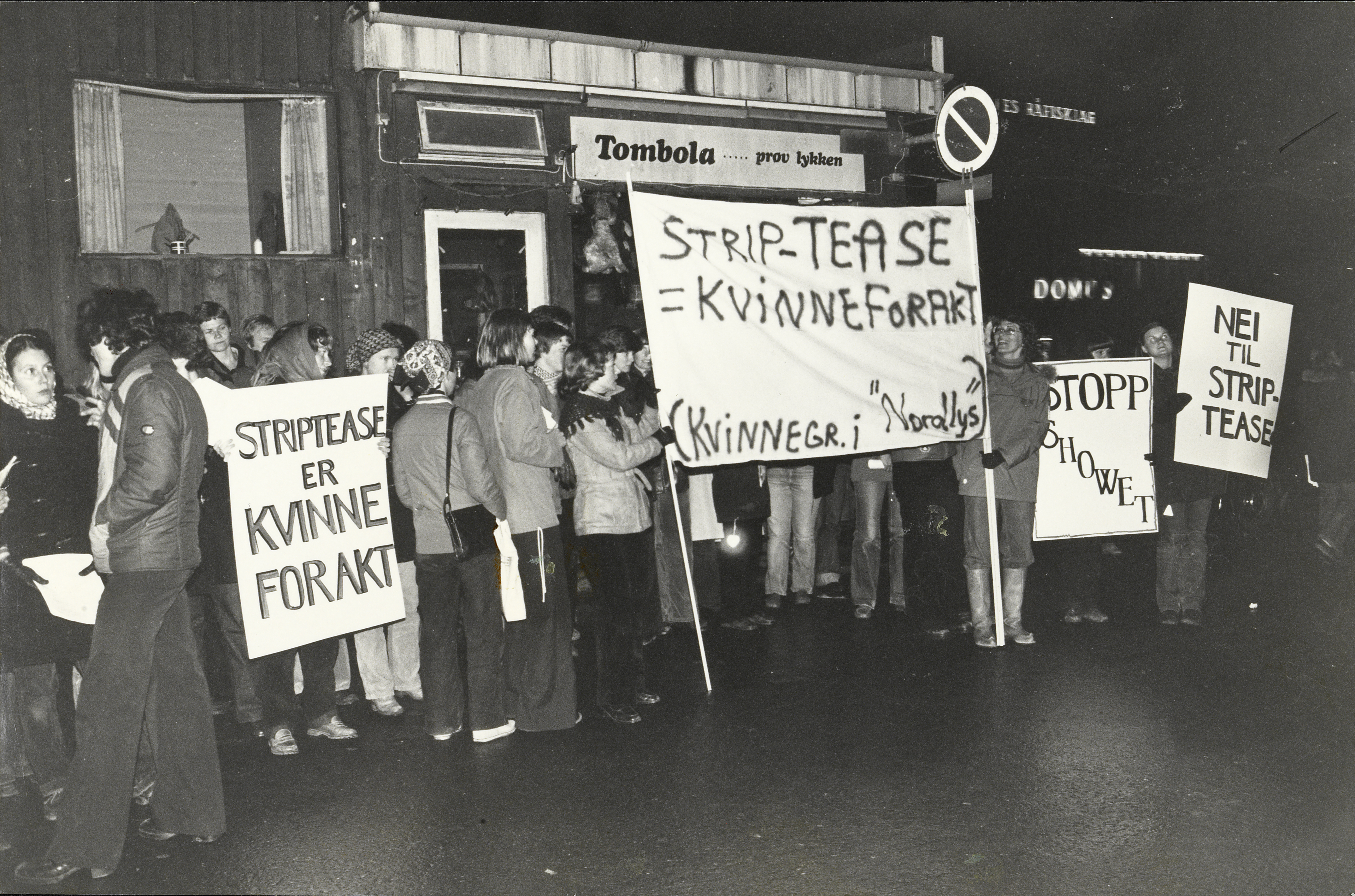 Demonstrasjon foran bygning "Tombola…. Prøv lykken" i Tromsø. Norges Råfisklag i bakgrunnen.  Kvinnelike demonstranter med plakater og banner med Striptease er kvinneforakt. Stopp showet. Nei til strip-tease (kvinnegr. i "Nordlys").