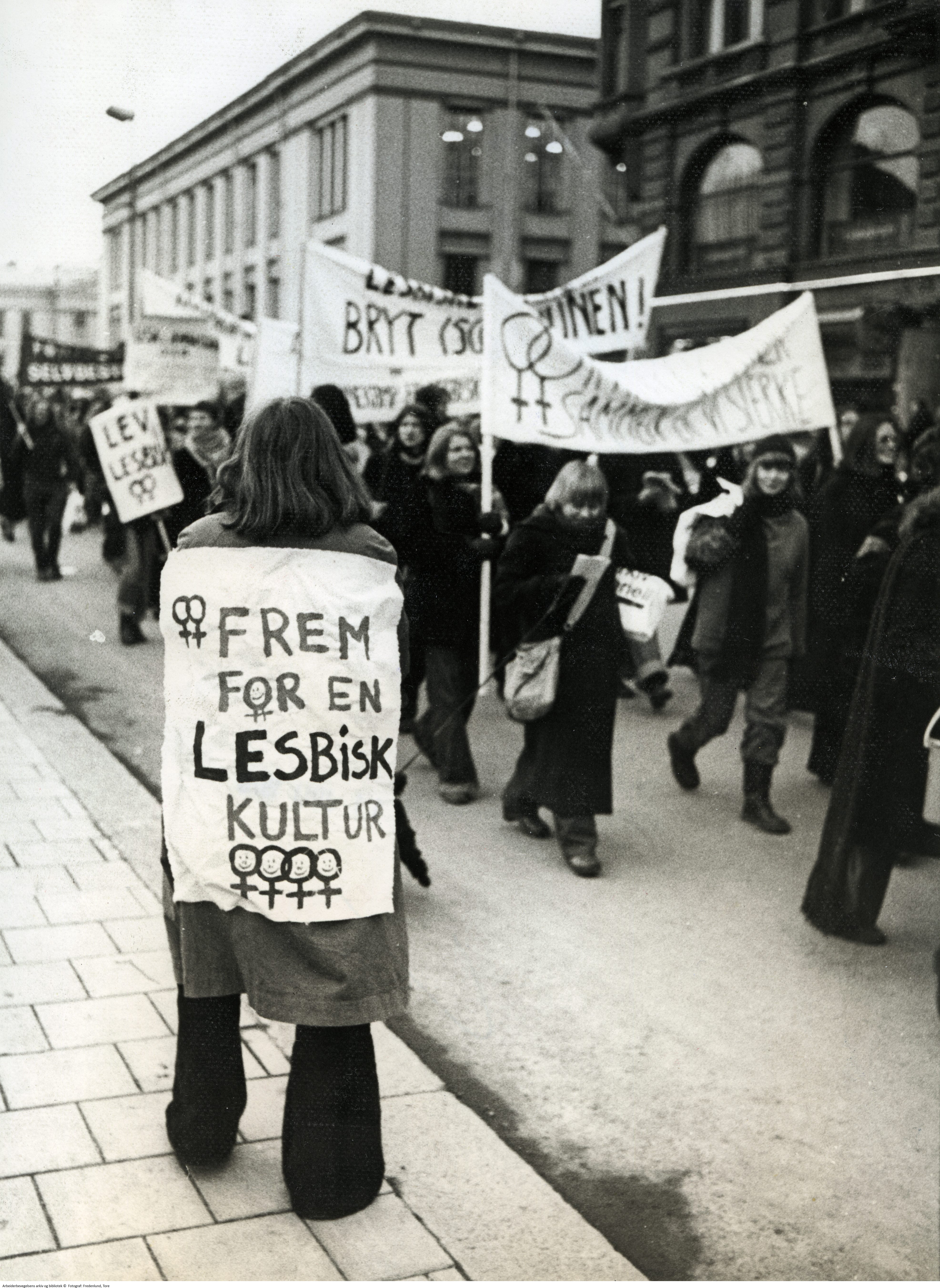 Demonstrasjonstog på Karl Johan i Oslo med paroler og plakater som "Lev lesbisk". I forgrunnen en kvinne med parole på ryggen: "Frem for en lesbisk kultur" dekorert med kvinnetegn.