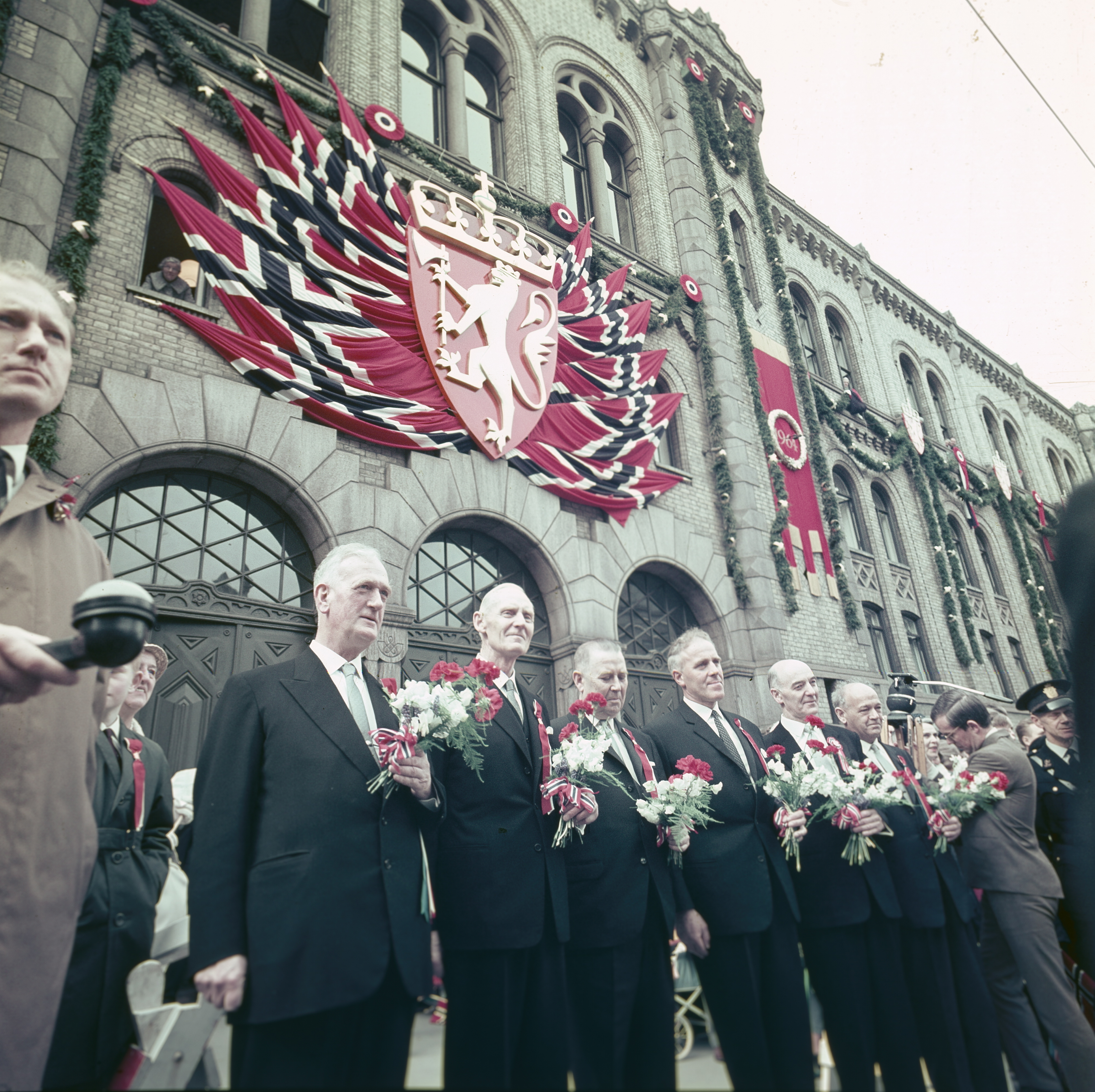 Gruppe menn med blomster i rødt, hvitt og blått står foran et pyntet Storting i forbindelse med Grunnlovsjubileet i 1964.